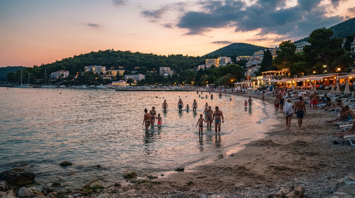 Uvala Lapad at sunset, shallow calm water, families wading far from shore and beach bars glowing along the pebble-and-sand crescent